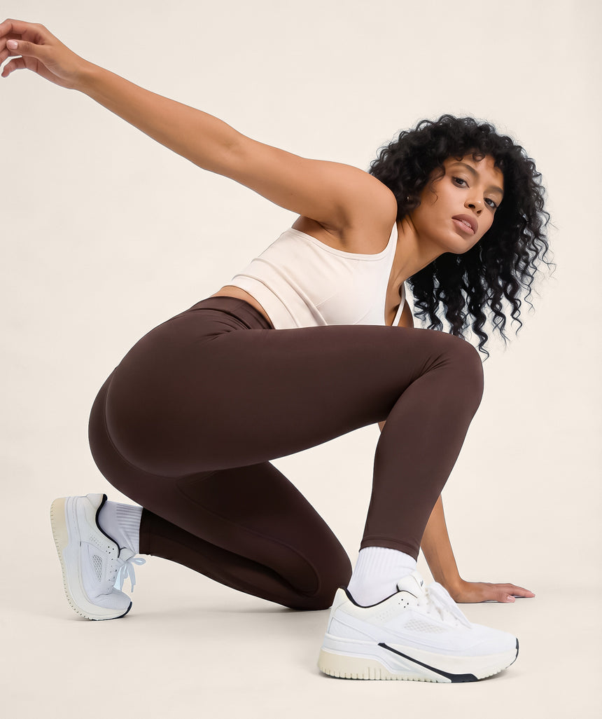 Woman in coffee brown leggings and white sneakers posing against a beige background