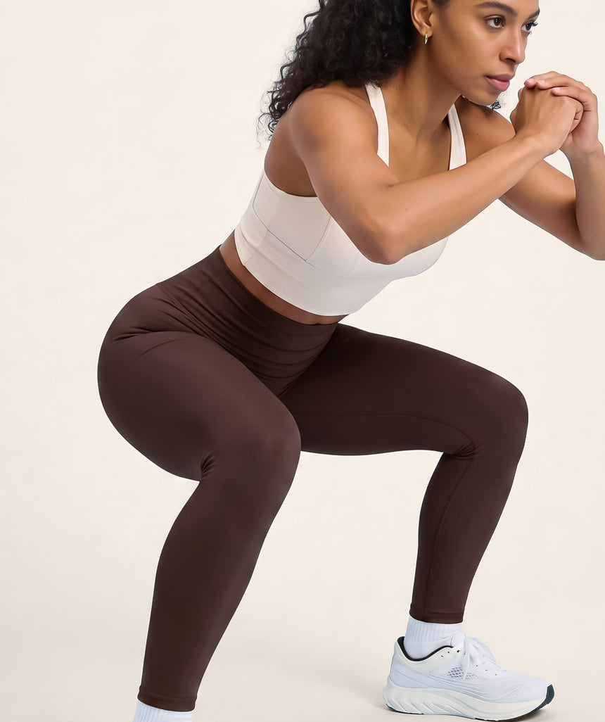 Woman in white sports bra and coffee brown leggings performing a squat on a beige background