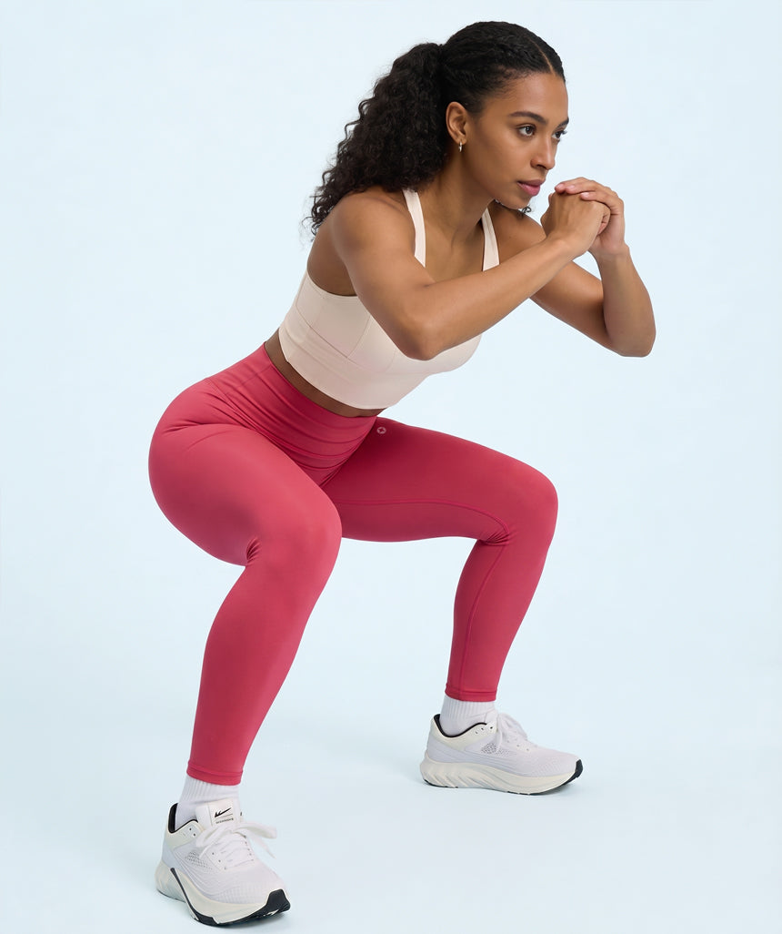 Woman in raspberry red leggings and white sports bra performing a squat on a light blue background